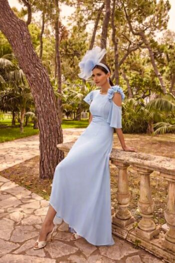 Woman in a pale blue gown with a large fascinator posing outdoors beside a stone balustrade in a garden park setting.