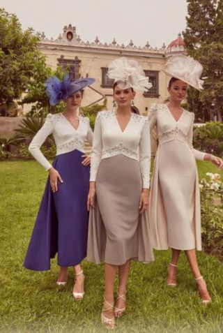 Three women posing in a garden in elegant midi dresses with large hats, in white and pastel tones, in front of a historic building.