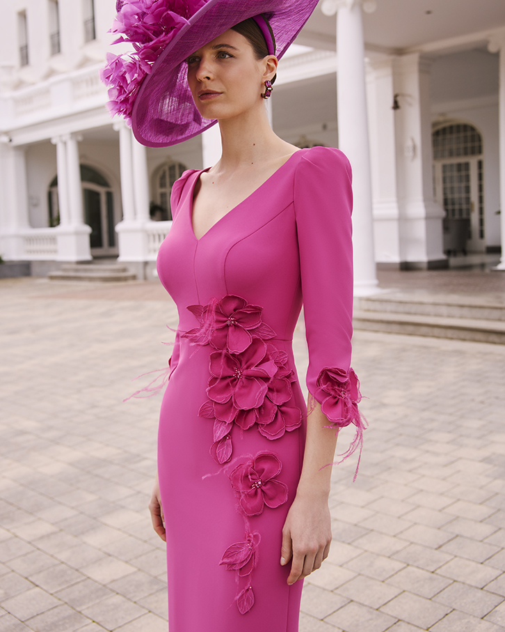1GL4 1 - Sofi Designs Bridal Woman in a hot pink dress with fabric flower embellishments and a matching wide-brim hat, standing outdoors near a white building with columns.