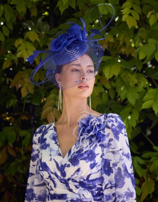 1GN7 3 - Sofi Designs Bridal Woman wearing a blue floral dress and an ornate blue fascinator with netting, posing outdoors against greenery.