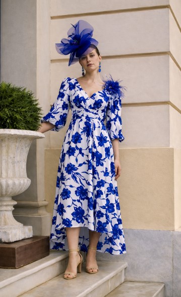 1GN7 4 - Sofi Designs Bridal Woman in a white dress with blue floral pattern, beige sandals, and a large blue fascinator standing on steps by a planter.