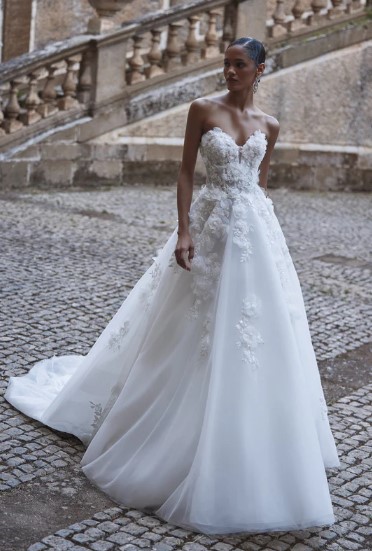 AADHYA 7 - Sofi Designs Bridal Bride in a strapless white wedding gown with lace appliqués, standing on a cobblestone surface with a stone staircase in the background.