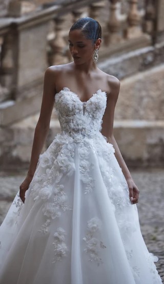 AADHYA 8 - Sofi Designs Bridal Bride wearing a strapless white wedding gown with floral appliqué bodice and full skirt, standing outdoors by stone architecture.