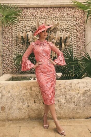 Woman in a pink lace sheath dress and matching pink hat posing with hands on hips in front of a stone fountain and tropical plants.