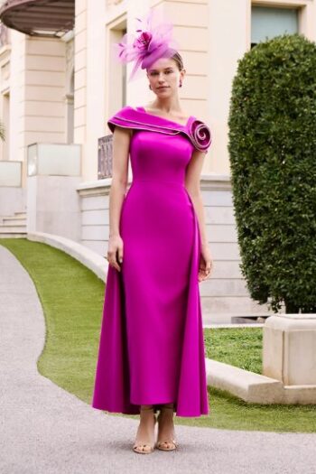 Woman in a magenta evening gown with a sculpted shoulder detail and pink feather fascinator standing outside a cream building.