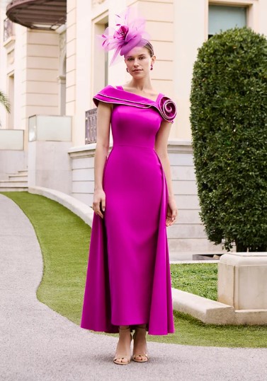 1G88 6 - Sofi Designs Bridal Woman in a magenta evening gown with a sculpted shoulder detail and pink feather fascinator standing outside a cream building.