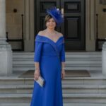 Svajone Kuzminskiene - Sofi Designs Bridal Woman in a royal blue off-shoulder gown with a matching blue fascinator, holding a clutch on stone steps before a dark door.