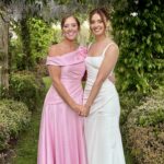 1 - Sofi Designs Bridal Two women in formal dresses posing together in a garden, one in a pink off-the-shoulder gown and the other in a white dress, smiling under a leafy archway.