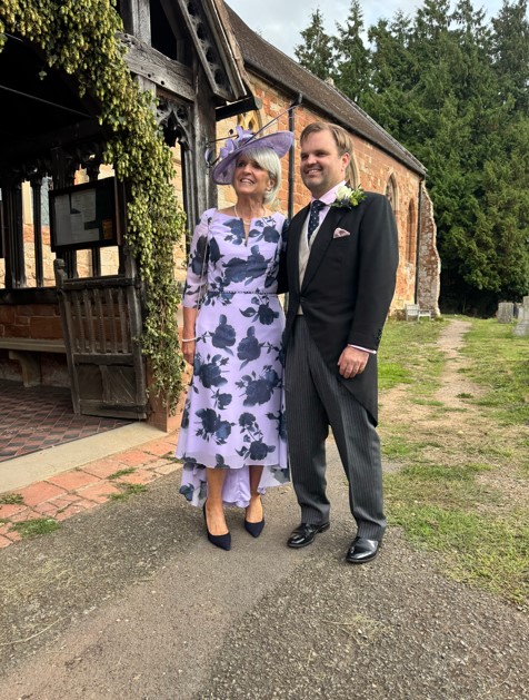 574572405_1489419575924748_2678213027319383297_n - Sofi Designs Bridal A couple in formal wedding attire stands outside a rustic brick building: woman in a lavender floral dress with a matching hat, and a man in a black morning coat with a boutonniere, smiling on a gravel path.