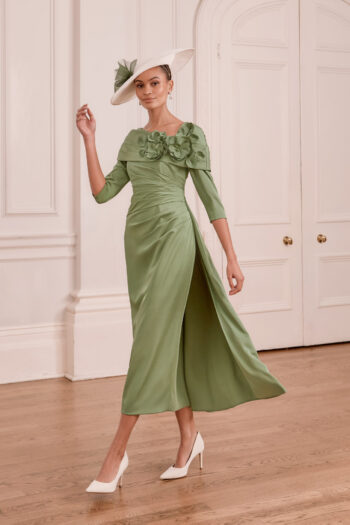 Elegant woman in a sage-green dress with floral appliqué, paired with a white hat, posing in a white paneled room with arched doorways.