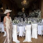 Screenshot - Sofi Designs Bridal Smiling woman in a champagne gown with lace and a large hat stands beside a white-clothed table at an elegant wedding reception with chandeliers in the background.