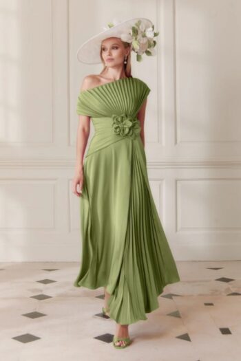 Woman in a light green, one-shoulder pleated gown with a floral waist corsage, paired with a white wide-brim hat adorned with flowers, standing indoors.