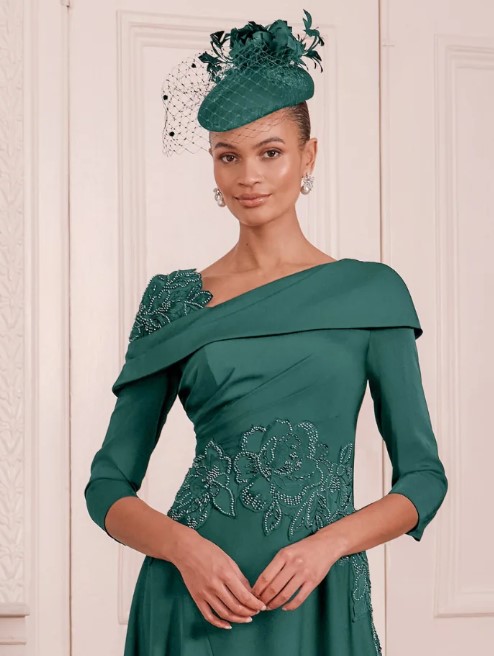 66797 Ivy 2 - Sofi Designs Bridal Elegant woman in a teal, off-the-shoulder gown with floral beading, wearing a matching teal hat with veil indoors.