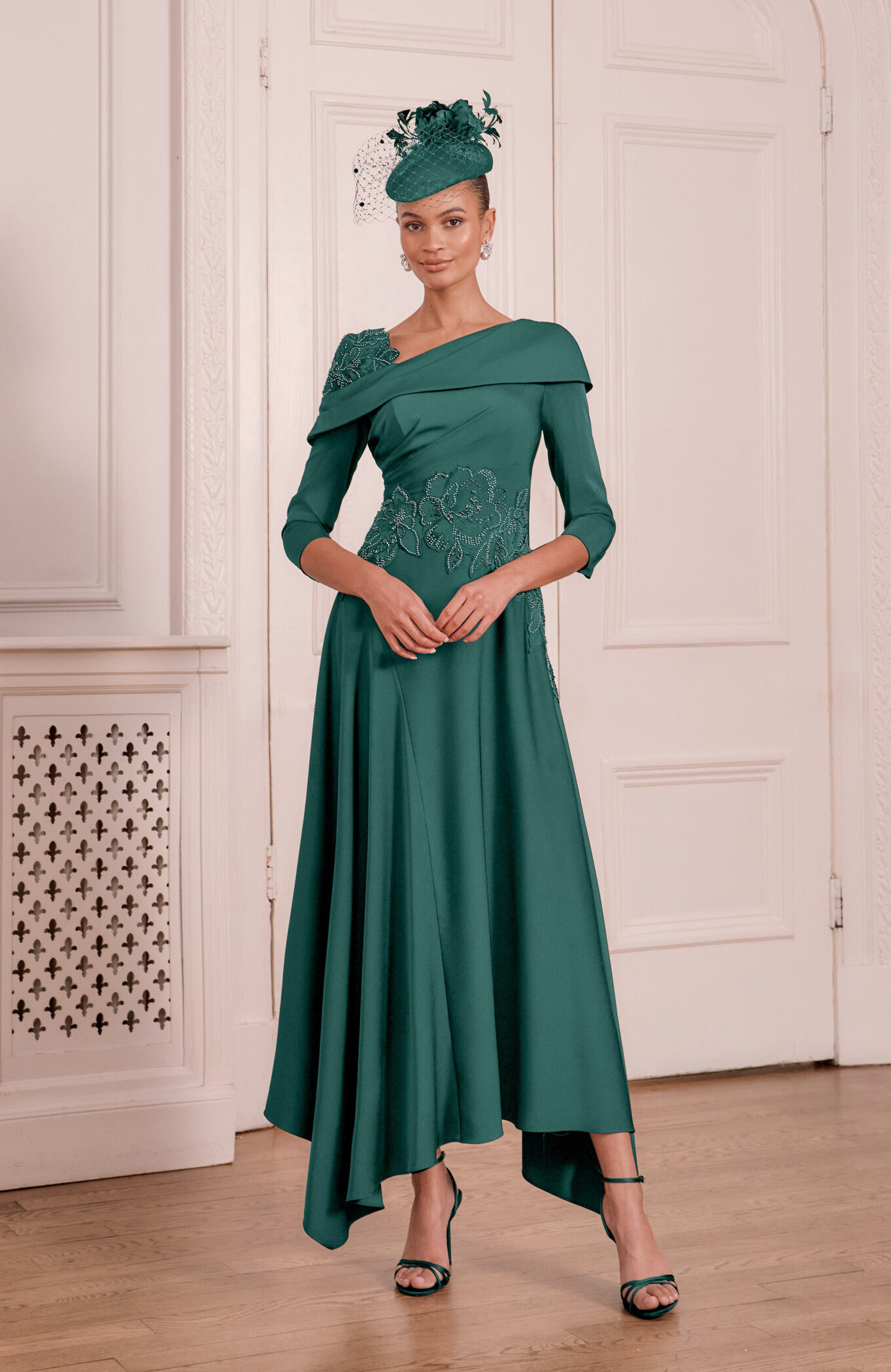 66797 Ivy - Sofi Designs Bridal Elegant woman in a teal off-shoulder gown with lace details and a teal fascinator, standing in a white ornate room.