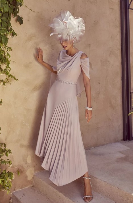 992422 1 - Sofi Designs Bridal Woman in a pale pink, off-shoulder dress with a pleated asymmetric skirt posing against a beige wall, wearing a large white fascinator hat and silver heels.