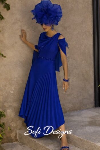 Woman in a royal blue, pleated dress with shoulder cutouts and matching oversized fascinator hat, standing against a beige wall and wearing blue sandals.