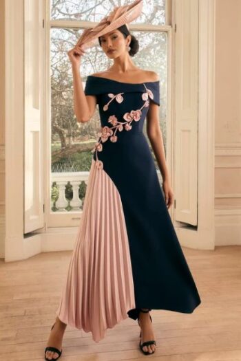 Woman wearing an off‑the‑shoulder navy evening gown with a pink pleated panel and floral embroidery, plus a pink hat and black sandals indoors by a window.