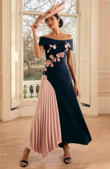 992814 Navy blush - Sofi Designs Bridal Woman wearing an off‑the‑shoulder navy evening gown with a pink pleated panel and floral embroidery, plus a pink hat and black sandals indoors by a window.
