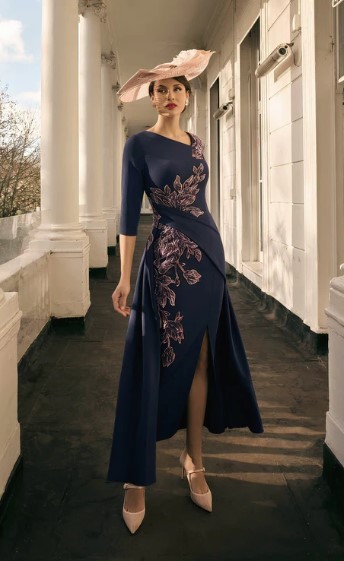992816 1 - Sofi Designs Bridal Woman in a navy dress with pink floral embroidery, deep side slit, cream heels, and a wide pink hat, standing on a sunlit columned balcony.