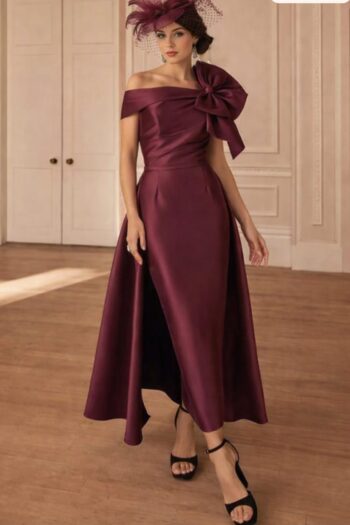 Woman in a burgundy satin off-shoulder gown with a large bow at the waist, wearing a matching fascinator, standing in an elegant room.