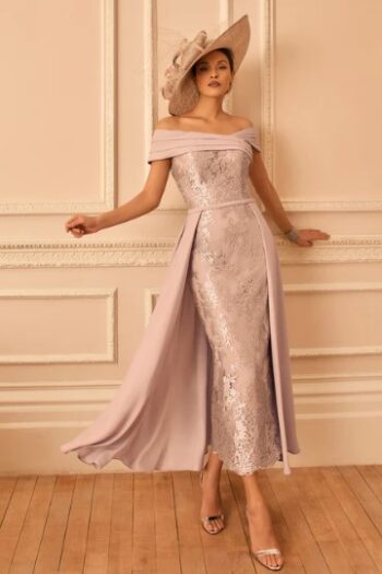 Model in a lavender off-shoulder sequin gown with a flowing cape and a wide-brim hat, posing by a decorative wall.
