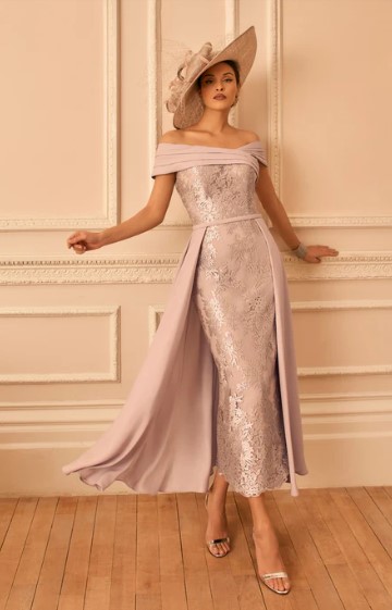 992842 Blush - Sofi Designs Bridal Model in a lavender off-shoulder sequin gown with a flowing cape and a wide-brim hat, posing by a decorative wall.