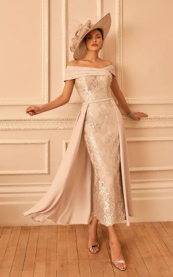 992842 Taupe 1 - Sofi Designs Bridal Woman in a pale pink off-the-shoulder lace dress with flowing cape panels and a wide-brim hat, posing by a paneled wall.