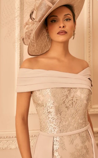 992842 Taupe 2 - Sofi Designs Bridal A woman in an off-the-shoulder beige lace gown with a pale pink belt and a large beige hat, standing against a decorative cream wall.