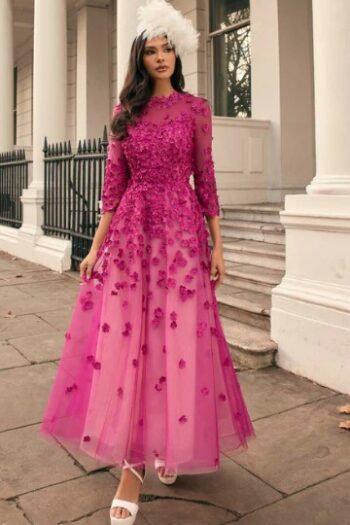 Woman in a magenta floral-embellished tulle gown with sheer sleeves and a white feather headpiece, posing on the steps of a white-columned building.