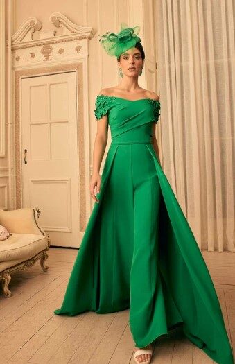 A woman wearing an emerald green off‑the‑shoulder gown with a dramatic pleated skirt and matching green fascinator, standing in an elegant, light-colored room.