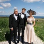 1 - Sofi Designs Bridal Three adults in formal wear stand on a gravel path with a grassy field and ocean in the background.