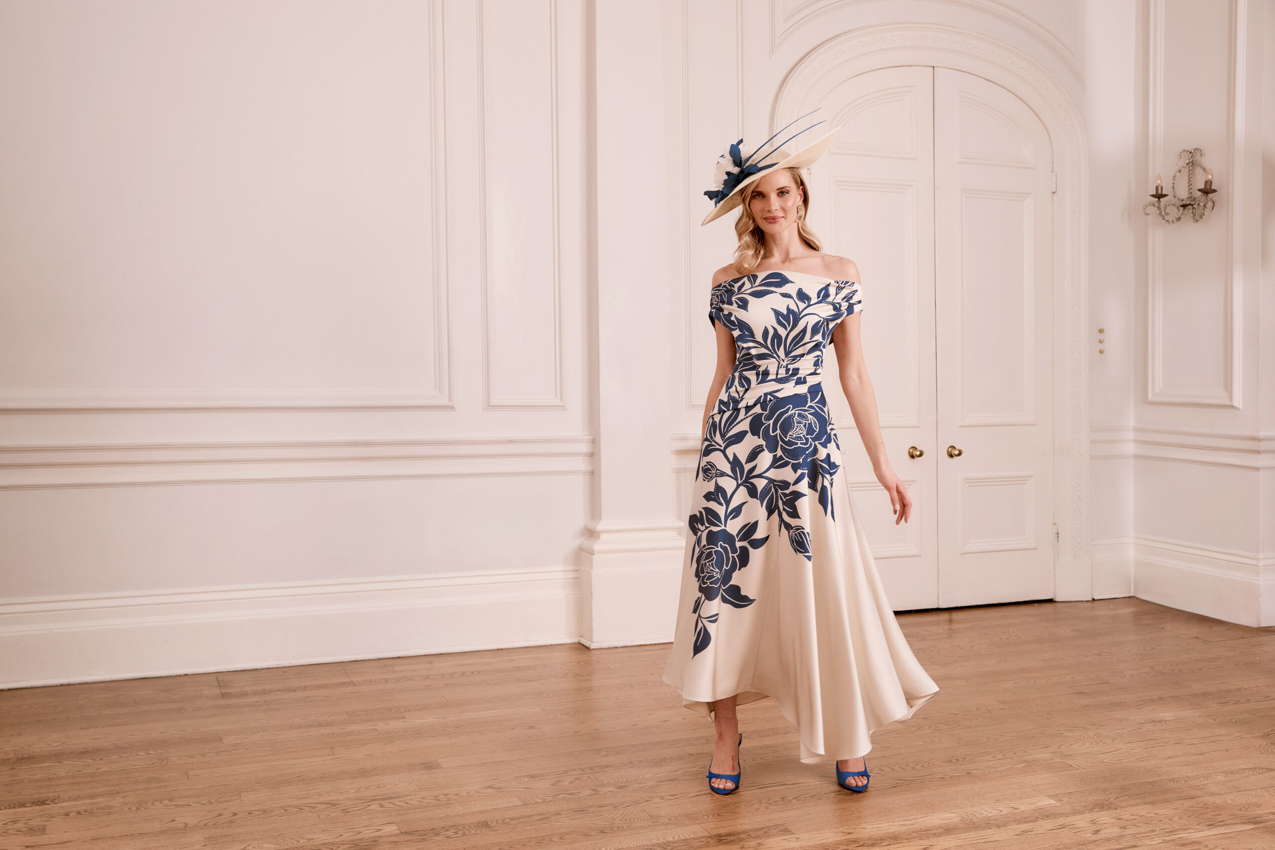 66780A 1 - Sofi Designs Bridal Woman in an off-the-shoulder cream gown with navy floral pattern, a blue hat, standing in a white paneled room with wood floor.