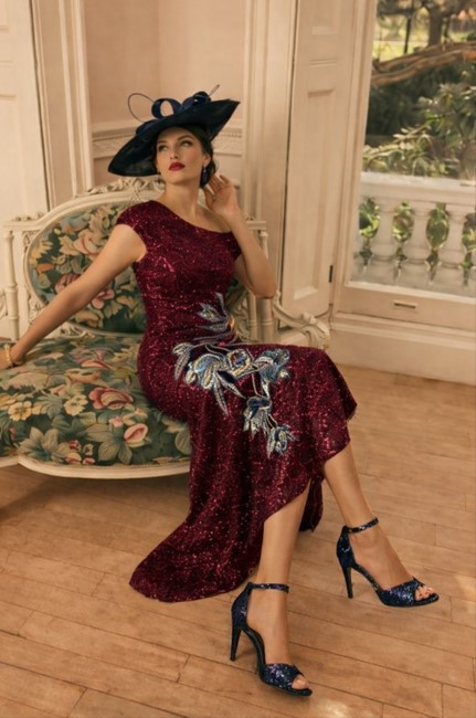 992803 Wine 2 - Sofi Designs Bridal Woman in a burgundy sequined gown and large black hat sits on a floral sofa in a bright, elegant room.