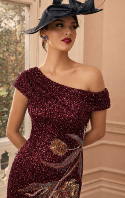 992803 Wine 3 - Sofi Designs Bridal Woman in a burgundy off-shoulder beaded gown with a decorative floral motif and a dark hat posing indoors by a window.