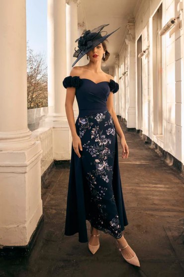 992809 Navy - Sofi Designs Bridal Woman in a navy off-shoulder evening gown with floral embroidery, puff sleeves, nude heels, and a large black fascinator hat, standing on a columned veranda.