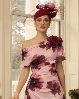 992827 Blush Wine print 2 - Sofi Designs Bridal Model wearing an off-shoulder pink floral dress with a large burgundy flower and a matching burgundy fascinator, indoors by a window.