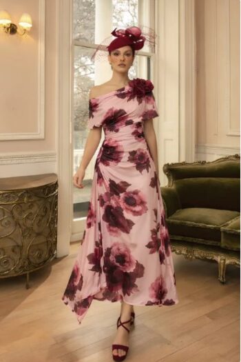Woman in a pink floral off-shoulder gown with large burgundy roses, wearing a matching burgundy fascinator, standing in an elegant room with a green sofa.