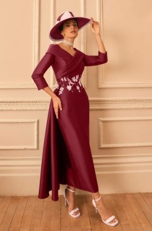 Woman wears a burgundy wrap-midi dress with floral embroidery, a matching wide-brim hat, white heels, standing against a beige paneled wall.
