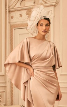 992847 Champagne 2 - Sofi Designs Bridal Model in champagne satin gown with draped wrap and cape sleeves, wearing a large white fascinator indoors.