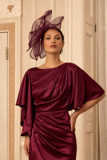992847 Wine 2 - Sofi Designs Bridal Woman in burgundy satin gown with draped bodice and cape sleeves, wearing a matching fascinator in an ornate hallway.