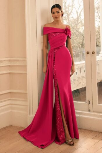 Woman in a bright pink off-shoulder evening gown with a thigh-high slit and sequined panel, standing by glass doors.