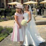 1 - Sofi Designs Bridal Bride in a white wedding gown holding a bouquet of white roses, posing with a woman in a pale pink dress and large hat outdoors
