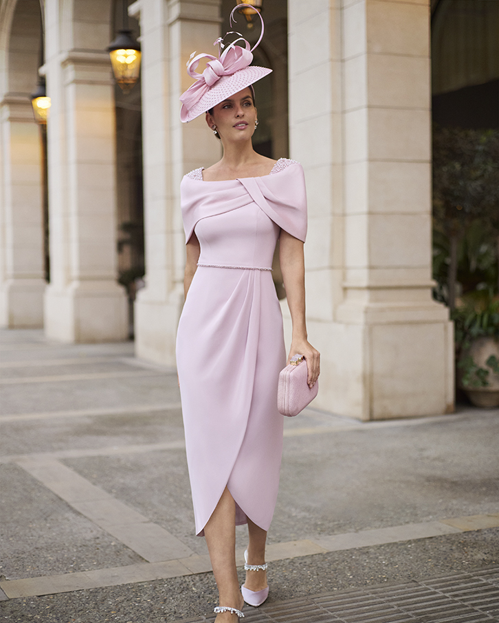 2G12_COUTURE_CLUB_3 - Sofi Designs Bridal Elegant woman in pale pink, off-shoulder wrap dress with a slim belt, matching pink hat with a bow, and a pink clutch, standing beside stone columns of a grand building.