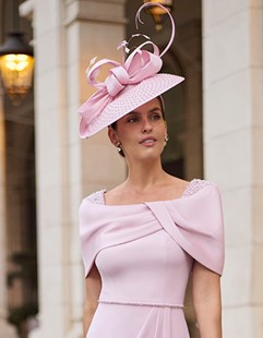 2G212 2 - Sofi Designs Bridal Woman posing outdoors in a pale pink, off-the-shoulder dress with a matching capelet and a large pink fascinator hat adorned with decorative spirals.
