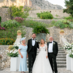 4 - Sofi Designs Bridal Bride in a white gown and groom in a tuxedo stand on stone steps with two parents, flanked by floral arrangements, against a garden wall and rocky hill backdrop.
