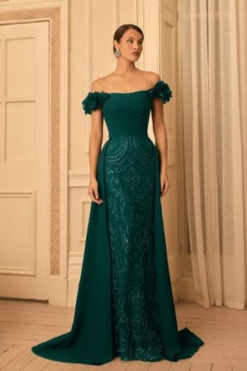 Woman in an emerald green off-the-shoulder evening gown with floral-ruffled sleeves, standing in a luxurious neutral room.