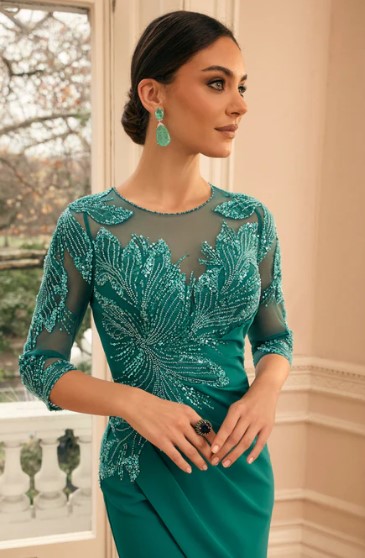 992875 2 - Sofi Designs Bridal Teal evening gown with beaded leaf embroidery and sheer sleeves; woman posing by a window with turquoise earrings.